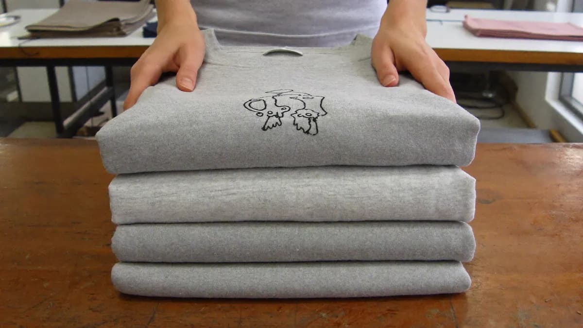 Pile de quatre pulls gris pliés sur une table d'atelier, le premier orné d'un petit motif brodé, mains posées dessus.